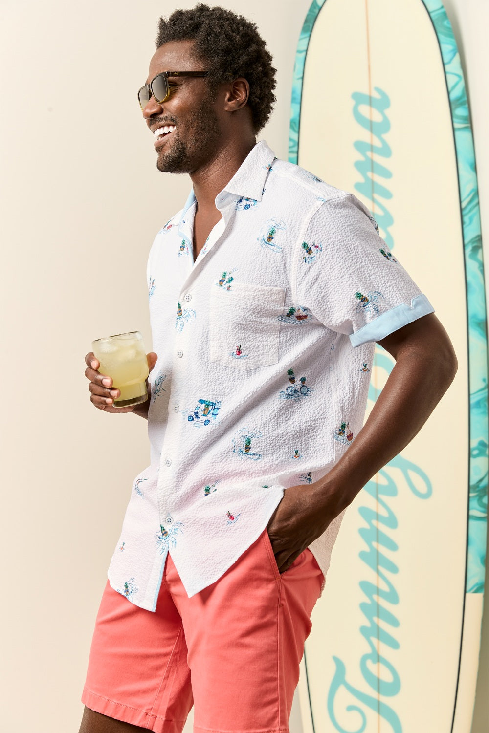 Man wearing a Tommy Bahama White shirt and pink shorts holding a drink, standing next to a surfboard with 'Tommy Bahama' branding.