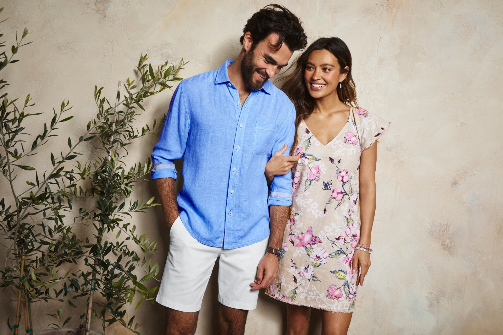 Man and woman standing together against a beige wall with plants.
