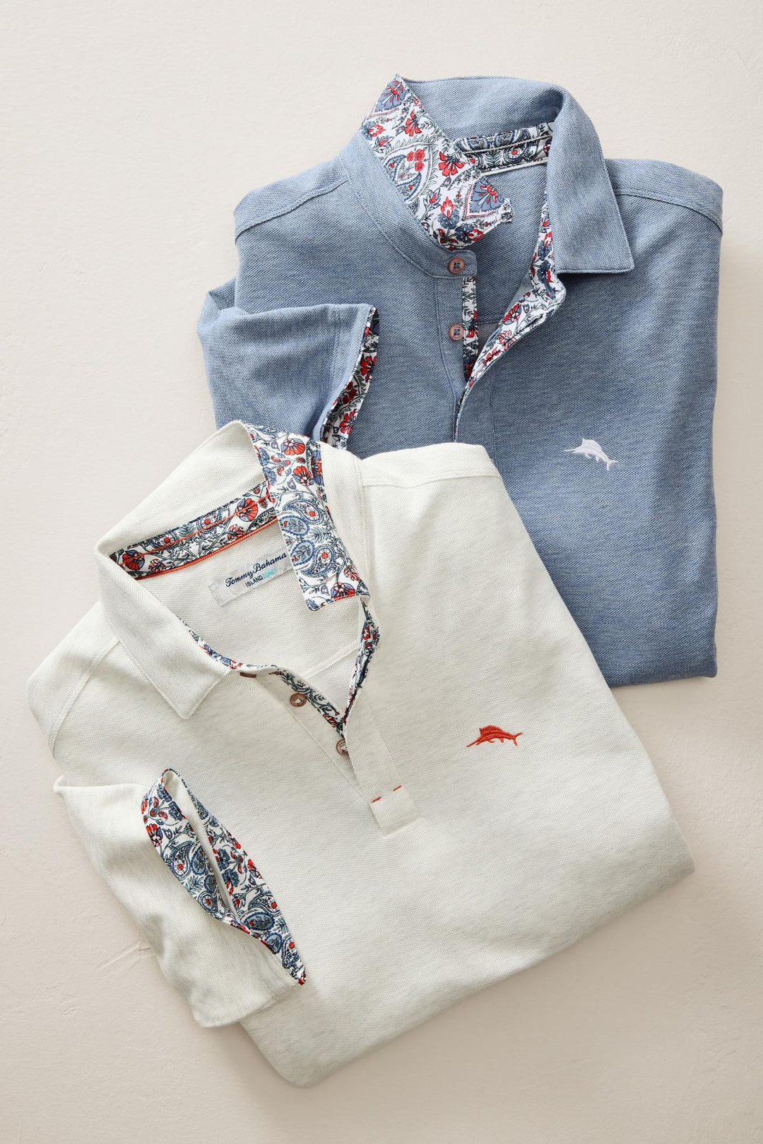 Folded Tommy Bahama blue and white polo shirts with floral collar details on a light beige background