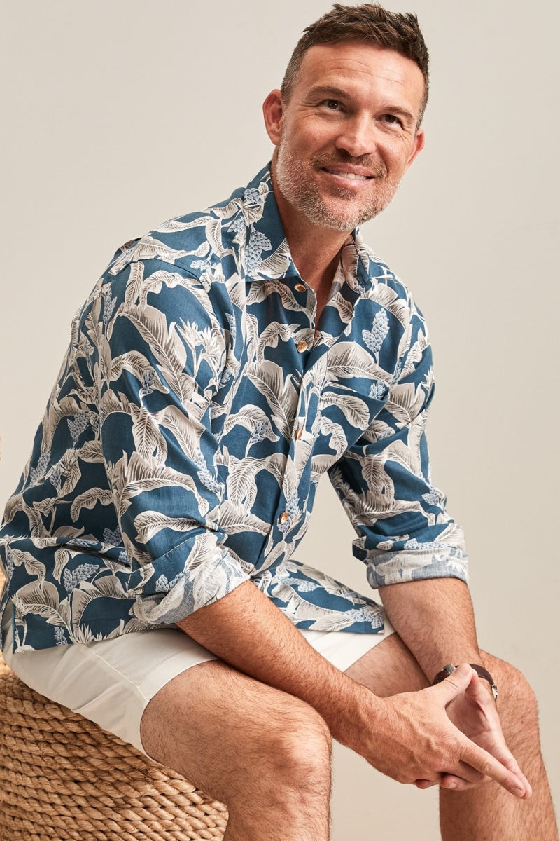 Man wearing a blue patterned shirt sitting on a woven chair against a plain background