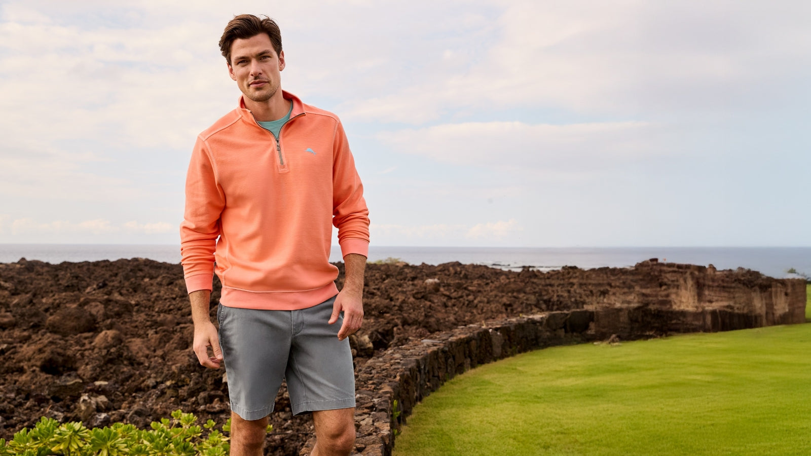 Man in Tommy Bahama peach pullover and gray shorts standing on a golf course with ocean view