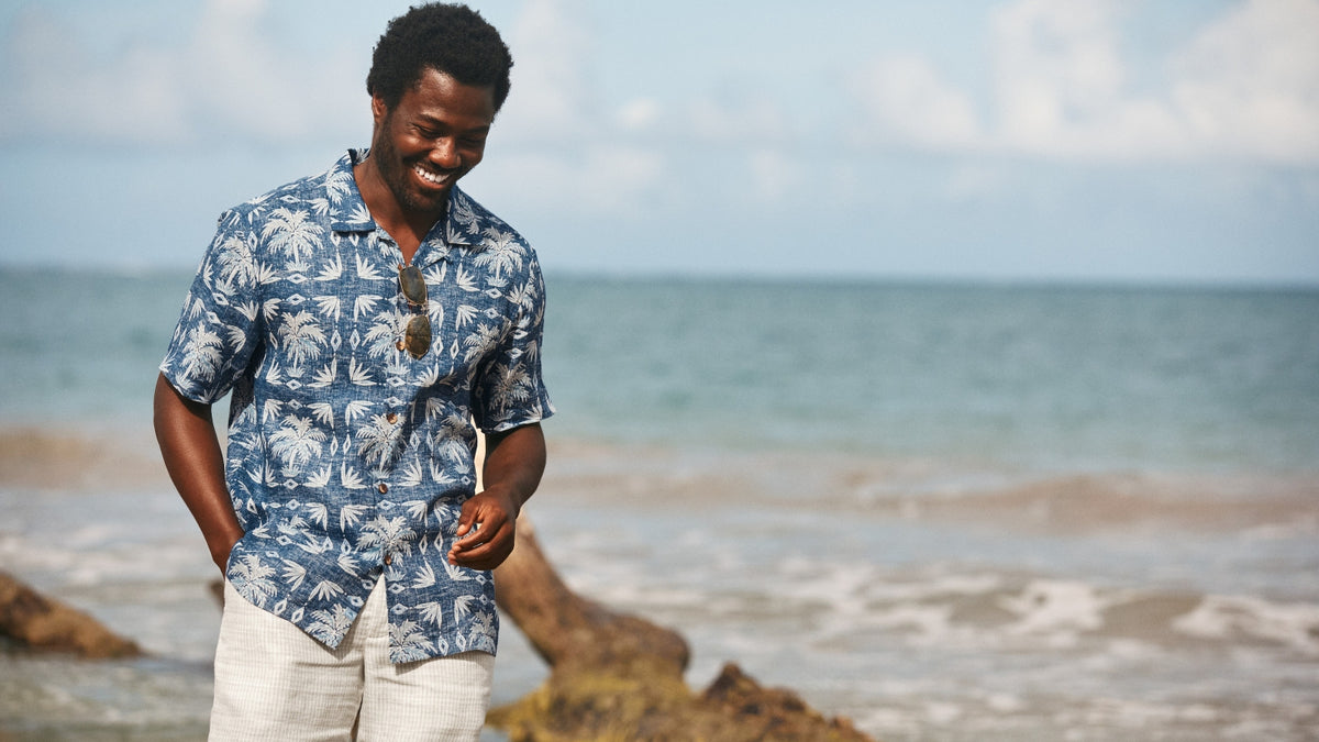 A man smiling on a beach wearing a blue and white palm tree patterned short-sleeve button-up shirt and white pants, with the ocean and sky in the background.