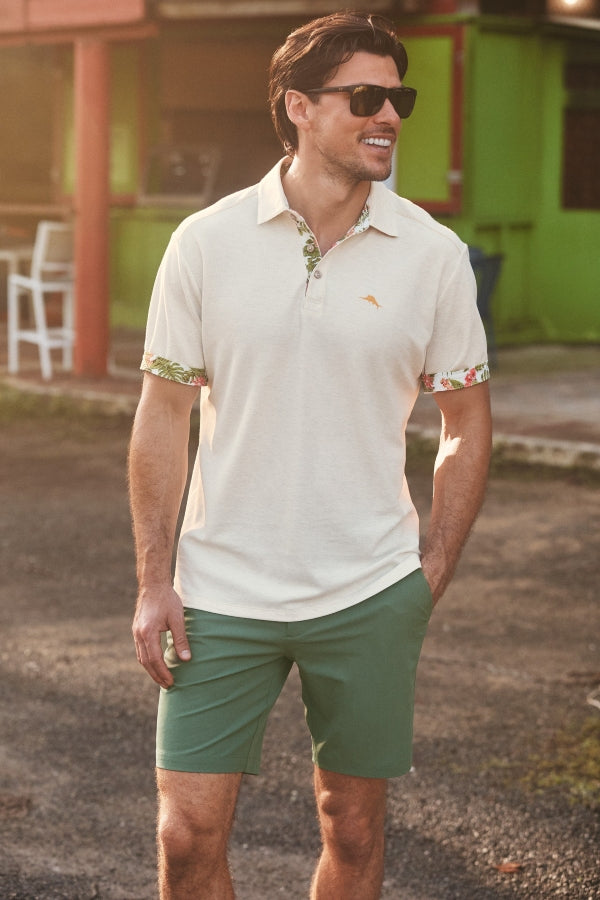 A man wearing a cream-colored short-sleeve polo shirt with green and white floral patterned cuffs, paired with green shorts, standing outdoors in front of a colorful building.