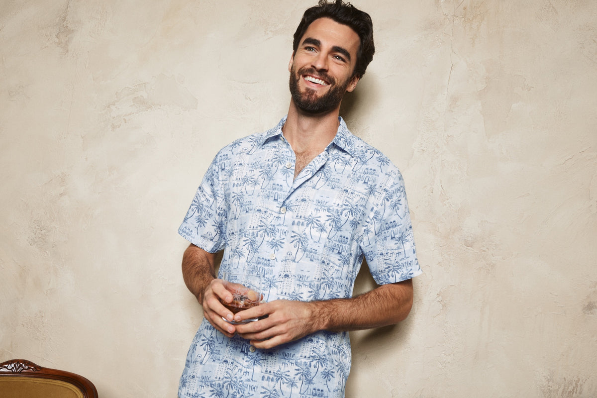 A smiling man with a beard wearing a light blue and white tropical palm tree print short-sleeve button-up shirt, standing against a textured beige plaster wall.